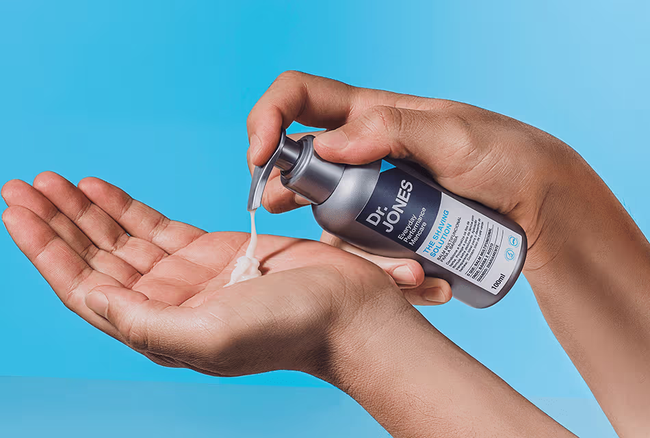 Foto mostra, em fundo azul, duas mãos. Uma delas aperta o pump do balm para barba The Shaving Solution, da Dr. JONES. A outra recebe o produto em sua palma.