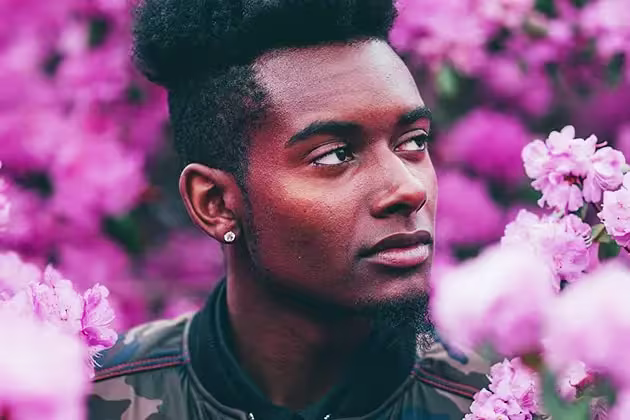 Em meio a campo de flores lilases, homem negro com cabelo raspado na lateral e alto no topo olha para a esquerda. Ele tem barba desenhada na lateral com cavanhaque bem longo, sem bigode e usa um brinco e jaquela de estilo militar.