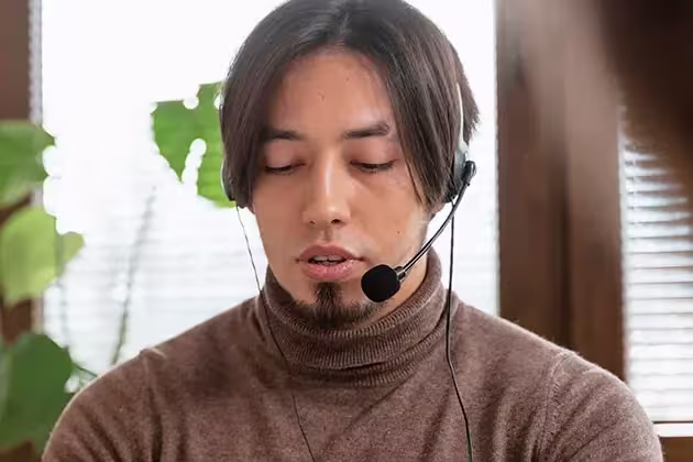 Homem oriental usa equipamento de telemarketing. Ele veste uma blusa cacharrel marrom e usa cavanhaque cheio sem bigode. Os cabelos são médios, castanhos e lisos.
