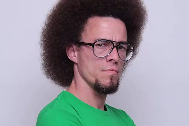 De camisa verde e óculos do tipo aviador, homem de pele clara e cavanhaque sem bigode olha para a câmera. Ele tem os cabelos crespos com estilo black power. A barba tem alguns grisalhos, e os cabelos são castanhos.