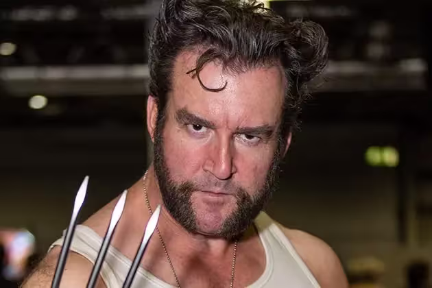 De camiseta branca, homem de pele clara imita Wolverine, com as garras do herói e cabelo e barba de mesmo estilo. A barba e os cabelos são castanhos.