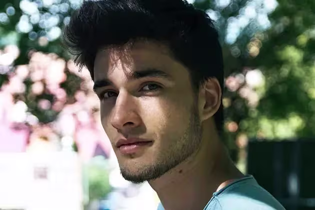 Jovem de pele clara e cabelos escuros com topete em frente a bosque olha para a câmera. Ele usa camiseta azul e tem a barba sem bigode e a fazer.