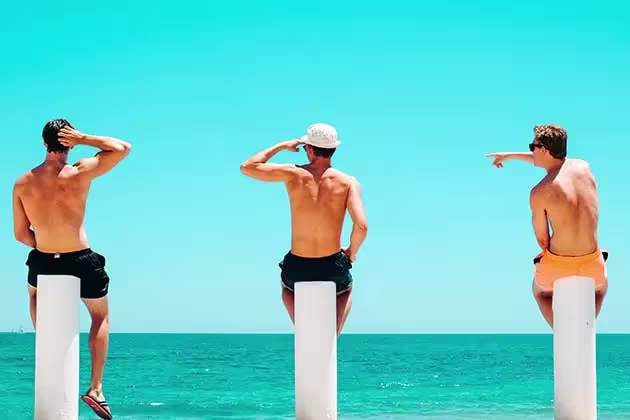Em frente ao mar, três homens brancos sem camisa estão de costas para a câmera, sentados em colunas brancas. Um usa boné branco (ao meio). Todos têm shorts (dois pretos e um laranja, à direita).