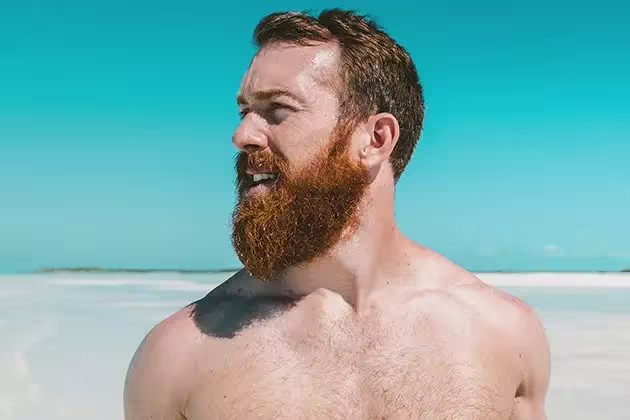 Em frente a praia deserta com muito sol, homem sem camisa e de pele clara, ruivo, olha para a esquerda. Ele tem a barba comprida, em alusão aos cuidados necessários para o verão.