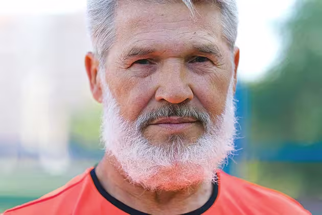 Foto mostra homem de cabelo grisalho, barba branca e pele clara em frente a paisagem, usando camiseta laranja de gola preta, olhando de frente para a câmera.