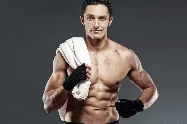 Em artigo sobre 6 séries de exercícios para fazer em casa, foto traz homem branco sem camisa, olhos claros e cabelos cacheados castanhos, toalha branca no ombro direito, luvas pretas para exercício, apontando para o abdômen.
