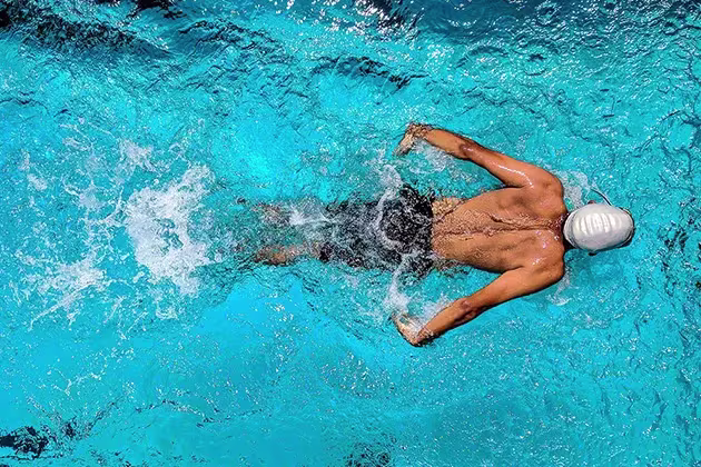Foto de homem de pele clara nadando em uma piscina azul, ao longe, com touca e traje masculino adequado para o esporte. A natação é uma das modalidades que permite queimar muitas calorias e ajudar no emagrecimento.