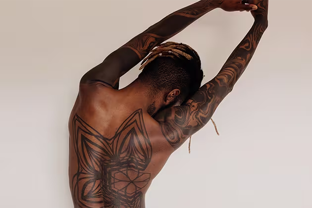 Homem negro, cabelo raspado na lateral, com tranças descoloridas. Ele está de costas, que são tatuadas, alongando-as, em frente a um fundo de cor clara, em artigo sobre série de exercícios para dor nas costas.
