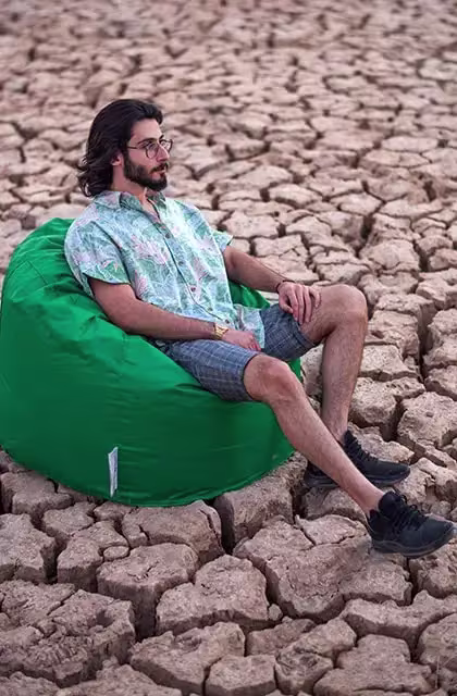 Foto de homem de ascendência árabe, de barba, óculos e cabelos compridos, sentado sobre um pufe, em meio a uma paisagem com um solo extremamente seco e rachado. Ele usa tênis preto, bermuda quadriculada azul-escura e camisa de mangas curtas azul-clara.