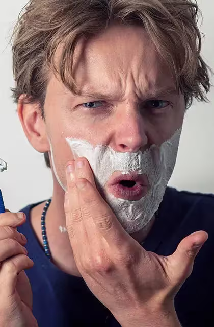 Foto de homem loiro, olhos azuis, com espuma de barbear no rosto. Ele usa camiseta azul, colar azul-claro e segura um aparelho de barbear, olhando direto para a câmera e fazendo careta, como se houvesse se cortado.