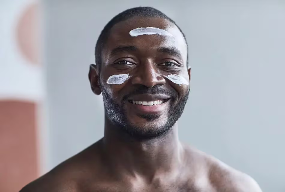 Foto de homem negro, cabelo raspado, barba curta, sorrindo para a câmera com resquícios de creme hidratante na testa e nas bochechas, com peito desnudo.