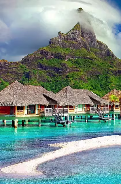 Foto de bangalôs sobre a água salgada em Bora Bora, com montanha ao fundo e com faixa de areia em destaque, à frente.