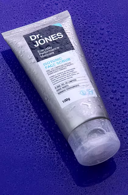 Em fundo roxo, com gotículas de água, foto em destaque da embalagem do gel esfoliante Isotonic Face Scrub, da Dr. JONES