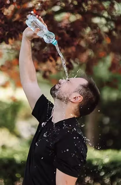 Homem de pele clara, cabelos curtos e barba joga água, a partir de uma garrafa, sobre o rosto. Ele está em um parque, e a imagem faz referência à hidratação.