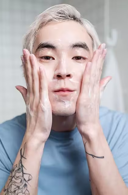 Foto de homem oriental, cabelos descoloridos, tatuagem nos braços, camiseta azul, passando gel de limpeza facial em frente à câmera.