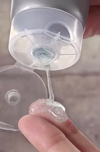 Foto de pessoa, de pele clara, colocando um pouco do esfoliante ISOTONIC FACE SCRUB, da Dr. JONES, em um dos dedos. A imagem mostra apenas as mãos e a embalagem do produto, em fundo neutro.