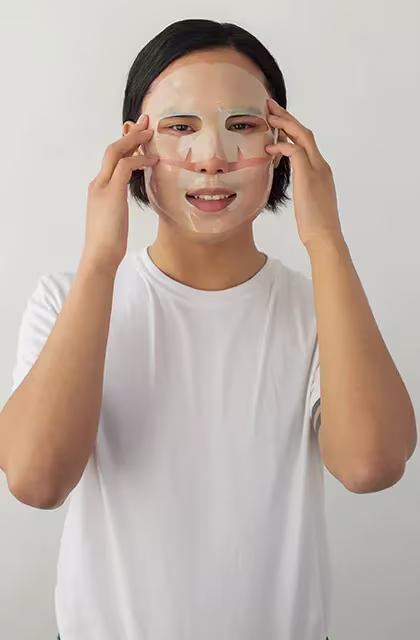 Em foto em fundo neutro, jovem asiático de cabelos escuros médios e camiseta branca põe uma máscara transparente no rosto, em alusão à pele sensível masculina.