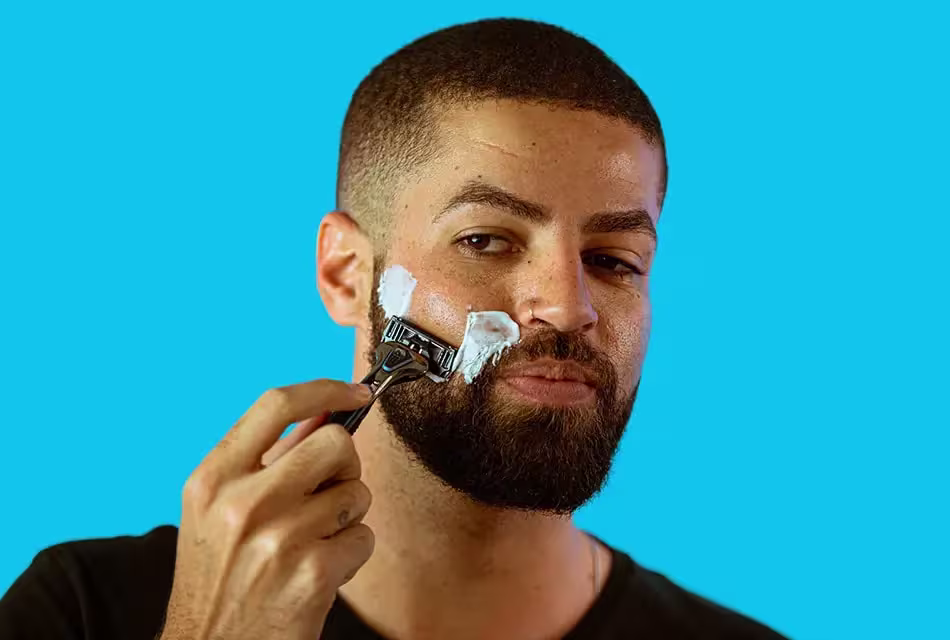 Em fundo azul, foto de homem de pele clara, camiseta preta, sardas e cabelo raspado fazendo a barba com a espuma de barbear PRECISION FOAM e com o aparelho THE RAZOR, da Dr. JONES.