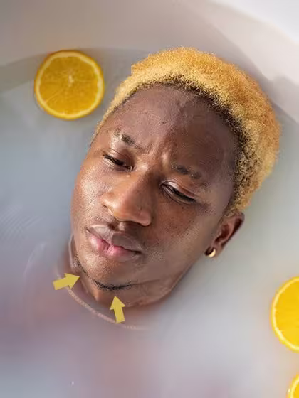 Foto de homem negro com cavanhaque ralo, sem bigode, deitado em banheira com sais de banho, cabelo colorido amarelo e rodelas de limão siciliano.