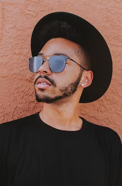 Foto de homem pardo, com barba aparada, cabelos e barba escuros, usando chapéu e camiseta preta em fundo laranja. A barba é fina nas laterais, com bigode e cavanhaque.