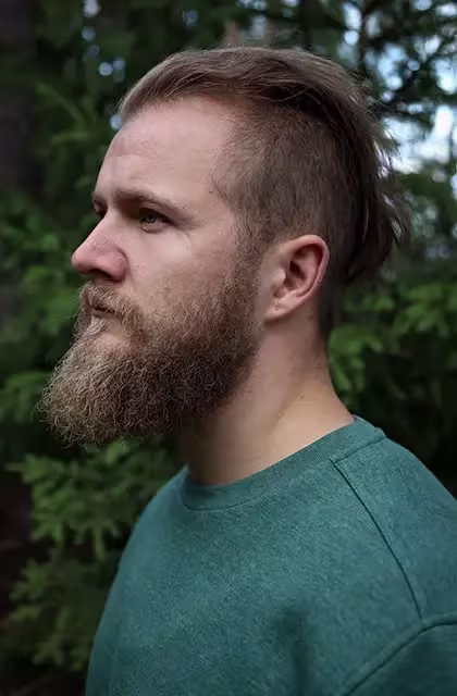 Foto de homem loiro, pele clara, cabelo 'sidecut' e barba estilo lenhador. Ele usa camiseta azul e está em frente a árvores, que estão ao fundo.