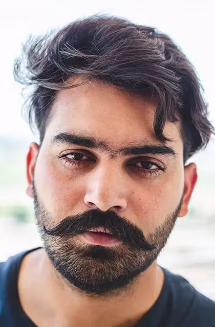 Foto de homem árabe, cabelos curtos, lisos e escuros e barba bem aparada no queixo e laterais e bigode volumoso estilo chevron. Ele está em frente a fundo neutro claro e usa camiseta azul-marinho.