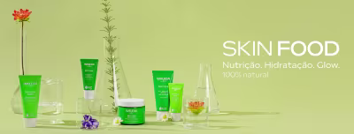 Linha Skin Food Weleda com fundo verde claro. Diversos produtos em tubos e potes verdes com rótulos Weleda Skin Food, acompanhados de flores naturais como rosa laranja, flor roxa e margarida branca, além de garrafas de vidro transparente. Texto em branco à direita: "SKIN FOOD Nutrição. Hidratação. Glow. 100% natural".