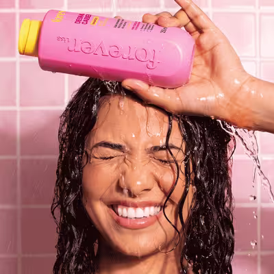 Mulher com cabelos molhados e pretos sorri enquanto despeja produto de frasco rosa com tampa amarela sobre sua cabeça.