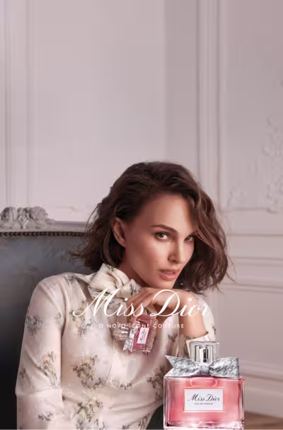 A imagem apresenta uma composição de flores rosas e brancas, com um frasco de perfume da marca Miss Dior no centro. O frasco possui um laço prateado e a embalagem exibe o nome da fragrância em destaque. Abaixo, há informações sobre uma oferta promocional que inclui uma amostra e uma miniatura do produto.