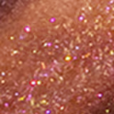 A imagem mostra a tampa da embalagem do gloss, com o aplicador virado pra cima, semelhante a uma tampa de batom, transparente e cilíndrica. Ao lado, o frasco do gloss, com o produto de cor marrom escuro e com a palavra KIND escrito na embalagem.