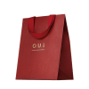 Sacola de presente vermelha em material texturizado, com alças e detalhes na mesma cor. Apresenta o logotipo "O.U.i" em letras douradas. A embalagem tem um design minimalista e elegante, com o vermelho vibrante como cor predominante.