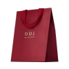 Sacola de presente vermelha em material texturizado, com alças e detalhes na mesma cor. Apresenta o logotipo "O.U.i" em letras douradas. A embalagem tem um design minimalista e elegante, com o vermelho vibrante como cor predominante.