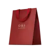Sacola de presente vermelha em material texturizado, com alças e detalhes na mesma cor. Apresenta o logotipo "O.U.i" em letras douradas. A embalagem tem um design minimalista e elegante, com o vermelho vibrante como cor predominante.