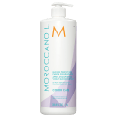 A imagem mostra o Condicionador Moroccanoil Color Care Blonde Perfecting Purple 1L, um frasco cilíndrico predominantemente branco, com detalhes em tons de roxo, laranja e azul. As informações do produto estão escritas em inglês e francês,