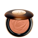 A imagem traz um sérum bronzeador compacto em uma embalagem. A tampa superior é marrom com alguns detalhes mais claros e tons mais escuros. O logotipo da marca aparece gravado em dourado no centro da tampa e o produto possui uma cor bege.