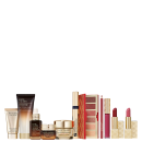 A imagem mostra uma variedade de produtos de beleza da marca Estée Lauder. Há frascos e embalagens de cosméticos e skincare, incluindo cremes, séruns, paleta de sombras, batons, gloss labial, delineador labial e máscara de cílios.