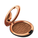 Pó bronzeador compacto em embalagem redonda de metal com acabamento dourado, contendo textura e padrão gravado na superfície do produto.