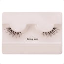 A imagem mostra os cílios postiços em um estojo plástico claro. O estojo tem uma cor nude claro e exibe um par de cílios postiços pretos.