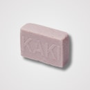 Barra de sabonete rosa claro em formato retangular. Tem textura levemente áspera e granulada. O nome "KAKI" está gravado em letras maiúsculas na superfície do sabonete. O tom rosado suave e a aparência rústica sugerem que pode ser um sabonete artesanal.