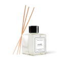 Difusor de aromas com varetas de madeira. Frasco transparente de vidro contendo líquido incolor. Rótulo preto e branco com o texto "arruda" e "Essential Organics". Três varetas longas de madeira clara inseridas no frasco para difundir a fragrância.