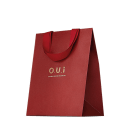 Sacola de presente vermelha em material texturizado, com alças e detalhes na mesma cor. Apresenta o logotipo "O.U.i" em letras douradas. A embalagem tem um design minimalista e elegante, com o vermelho vibrante como cor predominante.