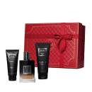 Conjunto. No centro, frasco de vidro contendo líquido da fragrância em degradê de preto à âmbar, de alto a baixo. Ao lado, tubo preto de gel de banho e um tubo preto de creme de barbear. Acompanha caixa vermelha com detalhes em dourado e laço vermelho.