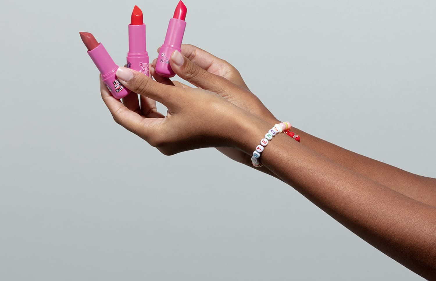 Três batons com embalagem e cores diferenes sendo segurados por mãos femininas negras contra fundo branco. No braço da modelo, vemos uma pulseira de miçangas brancas com detalhes coloridos.