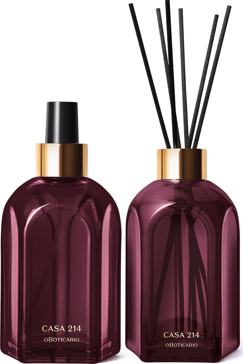 A imagem mostra dois vidros retangulares roxos, com rótulo em dourado. Um deles é o aromatizador de ambientes com válvula spray preta e o outro é um difusor com tampa dourada que acompanha varetas de madeira. Ambos são a linha Casa 214 Figo Magnífico.