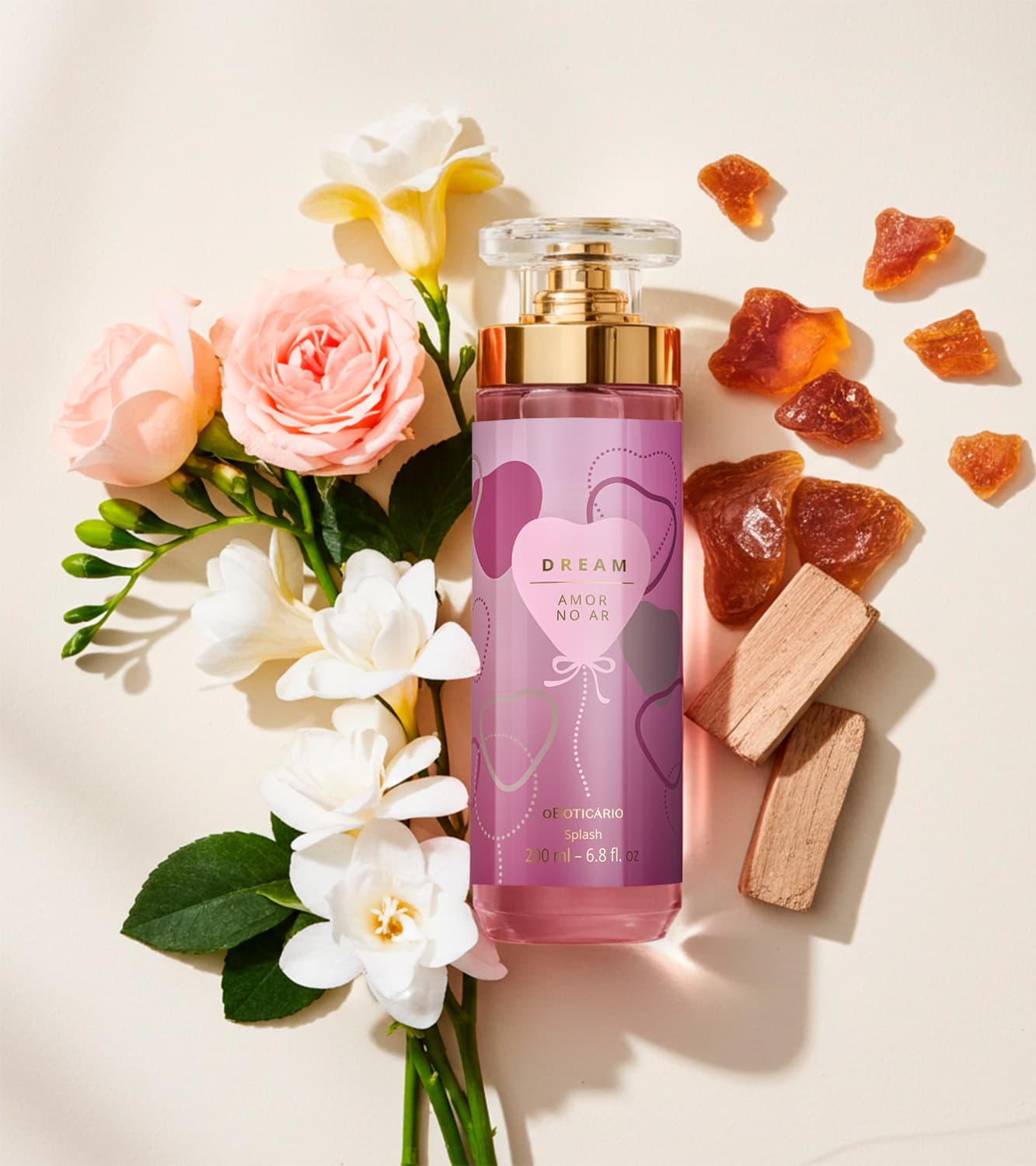 Imagem de um arranjo floral com rosas, orquídeas e folhagens verdes em fundo claro. Ao lado, frasco de body splash Dream Amor no Ar, com tampa transparente e válvula spray dourada. O rótulo é em tons de roxo e tem um coração estampado.