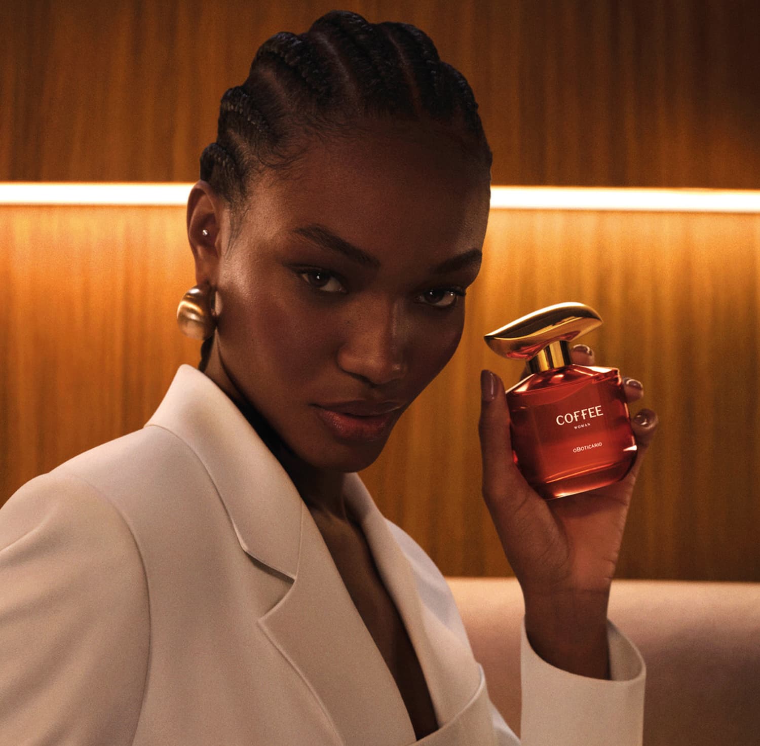 Imagem de uma mulher de pele negra segurando um frasco de desodorante colônia Coffee Woman Unique em um ambiente interno amadeirado com iluminação quente. Ela usa um casaco branco, brincos dourados e possui tranças no cabelo.