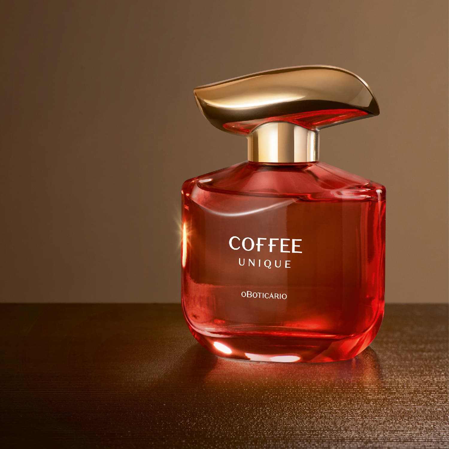 Imagem de um frasco de vidro avermelhado com tampa dourada de desodorante colônia da marca Coffee Woman Unique, em fundo marrom escuro, refletindo a luz em sua superfície brilhante e amadeirada.