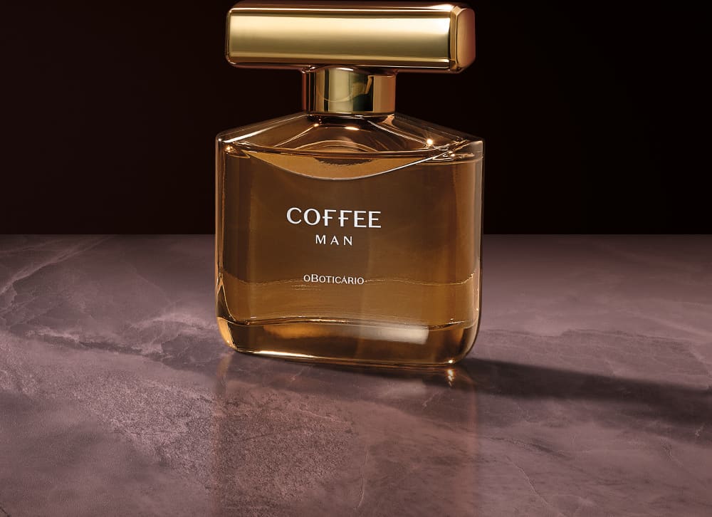 Imagem de um vidro do desodorante colônia Coffee Man em fundo escuro, com superfície de pedra e iluminação dourada. A embalagem possui uma luxuosa tampa com rotulo dourado.