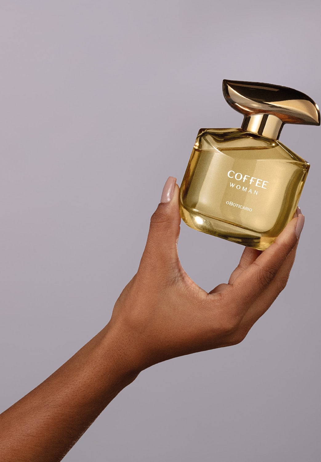 Imagem de um sofisticado frasco de desodorante colônia Coffee Woman Floriental Ambarado, com tampa dourada e cantos arredondados, sendo segurado por uma mão sobre fundo cinza claro, em cenário minimalista e elegante.