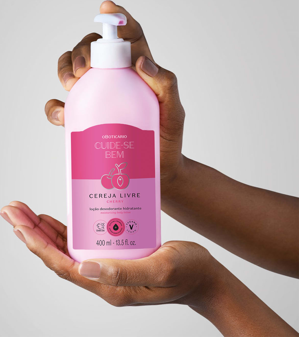Mão feminina segurando um frasco branco com bomba dosadora e rótulo nas cores rosa e lilás com cerejas ilustradas contendo loção hidratante corporal Cuide-se Bem Cereja Livre em fundo lilás claro.